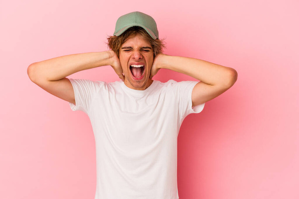 Homem caucasiano jovem com maquiagem isolada no fundo rosa cobrindo orelhas com as mãos tentando não ouvir som muito alto. - Foto, Imagem