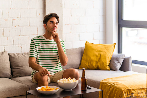 Joven hombre de raza mixta comiendo palomitas sentado en el sofá mirando de lado con expresión dudosa y escéptica. - Foto, Imagen