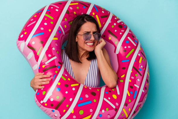 Young caucasian woman with inflatable donut isolated on blue background covering ears with hands. - Photo, Image