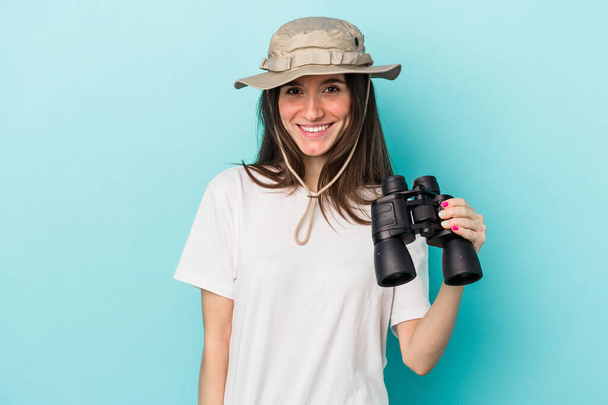 Young caucasian explorer woman holding binoculars isolated on blue background happy, smiling and cheerful. - Фото, зображення