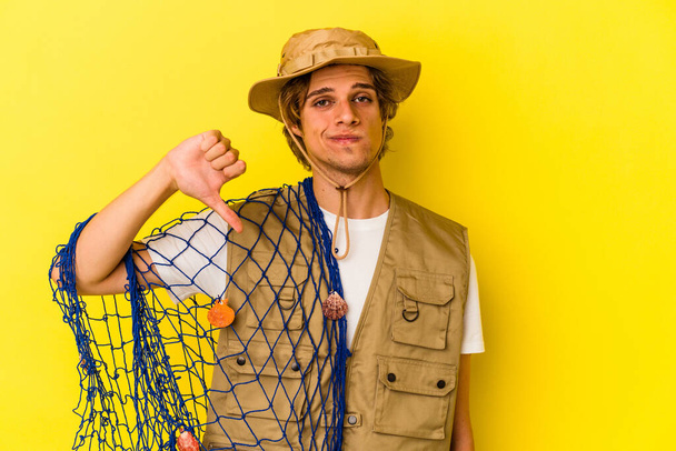 Mladý rybář s make-upem, který drží síť izolovanou na žlutém pozadí a ukazuje gesto nelibosti, sklopí palce. Koncept neshod. - Fotografie, Obrázek