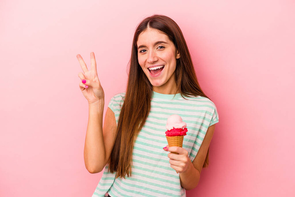Jovem caucasiana segurando um sorvete isolado em fundo rosa alegre e despreocupado mostrando um símbolo de paz com os dedos. - Foto, Imagem
