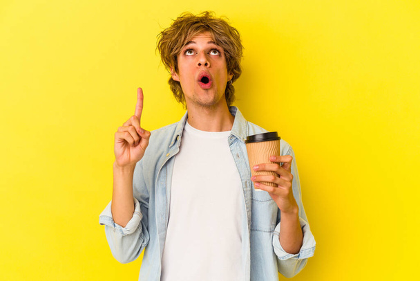Joven hombre caucásico con maquillaje sosteniendo un café para llevar aislado sobre fondo amarillo apuntando al revés con la boca abierta. - Foto, Imagen
