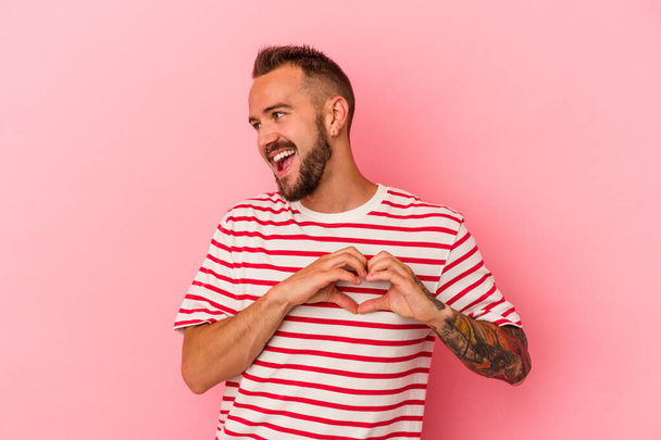 Jonge blanke man met tatoeages geïsoleerd op roze achtergrond glimlachend en met een hartvorm met handen. - Foto, afbeelding