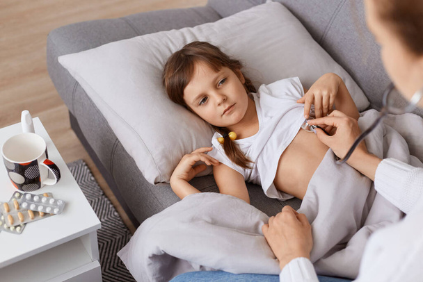 Caucasian little girl lying on pillow on the sofa, female doctor using stethoscope on a heartbeat or lungs at home, kid in white t shirt suffering headache and coughing. - 写真・画像