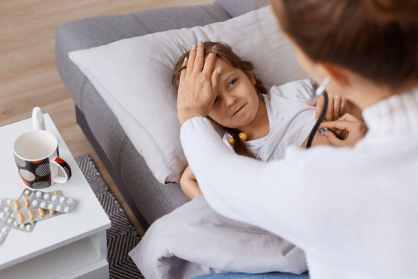 Внутренний снимок маленькой девочки, лежащей на диване, доктор, используя стетоскоп, чтобы осмотреть легкие детей, ребенок простудился во время холодной влажной осенней погоды, здравоохранение. - Фото, изображение