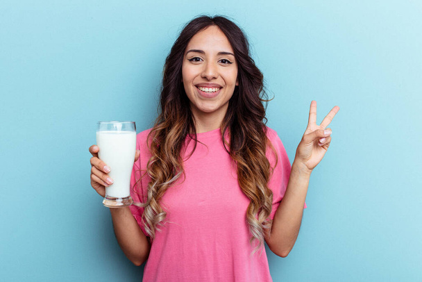 Mavi arka planda bir bardak süt tutan genç melez kadın neşeli ve kaygısız parmaklarıyla barış sembolü gösteriyor.. - Fotoğraf, Görsel