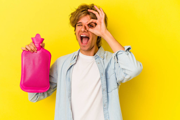 Young caucasian man with makeup holding a hot bag of water isolated on yellow background  excited keeping ok gesture on eye. - Photo, Image