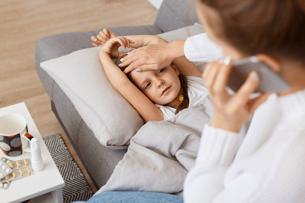 Side view of mother sitting near her unhealthy little child girl and calling a doctor because of her daughters sickness, asking about treatment, touching kid forehead. - Valokuva, kuva
