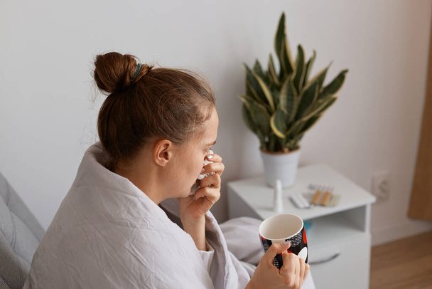 Sick woman with hair bun with headache sitting under the blanket with cup of tea or medicine, suffering runny nose, sore throat and coughing, female with influenza. - Foto, immagini