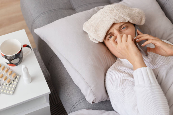 Unhealthy woman with towel on her forehead swearing white sweater lying on sofa and talking via smartphone with daughter, using napkin for runny nose. - Fotografie, Obrázek