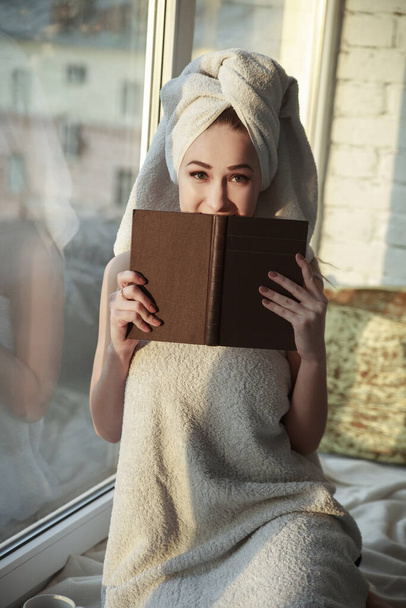 A cute girl in a towel is resting at the window on a sunny day with a book. - Fotografie, Obrázek