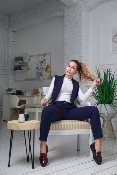 A cute girl in an office interior sits in casual clothes sits on a chair in full growth. - Foto, afbeelding