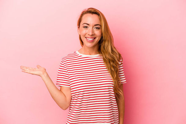 Caucasian blonde woman isolated on pink background showing a copy space on a palm and holding another hand on waist. - Photo, Image
