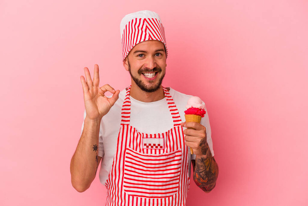 Nuori valkoihoinen jääpalakone mies tatuoinnit tilalla jäätelöä eristetty vaaleanpunainen tausta iloinen ja luottavainen osoittaa ok ele. - Valokuva, kuva