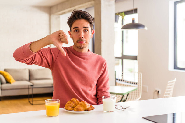 Fiatal kevert fajú férfi reggelizik a konyhában reggel, egy ellenszenves gesztust mutatva, hüvelykujjal lefelé. A nézeteltérés fogalma. - Fotó, kép