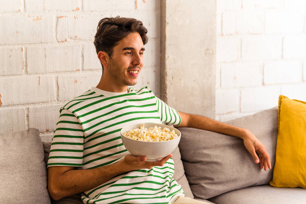 Jonge gemengde ras man eet popcorn zittend op de bank - Foto, afbeelding