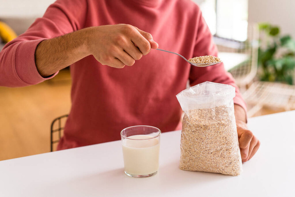 Young mixed race man eating oatmeal and milk for breakfast in his kitchen - Photo, Image