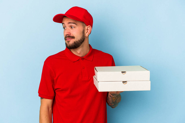 Fiatal fehér férfi tetovált tartja pizza elszigetelt kék háttér néz félre mosolygós, vidám és kellemes. - Fotó, kép