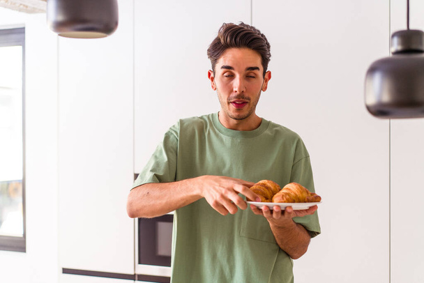 Jeune homme métis mangeant croissant dans une cuisine le matin - Photo, image
