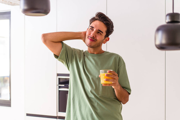Jovem mestiço bebendo suco de laranja em sua cozinha tocando na parte de trás da cabeça, pensando e fazendo uma escolha. - Foto, Imagem