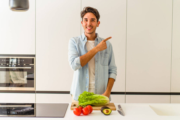 Jeune homme métis préparant une salade pour le déjeuner souriant et pointant du doigt, montrant quelque chose à blanc. - Photo, image