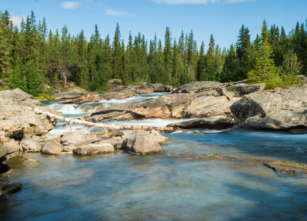 Prachtig landschap met lange blootstelling waterstroom en waterval van rivier de Kamajokk, rotsblokken en sparren boombos in Kvikkjokk dorp in Zweeds Lapland. Zomer zonnige dag, blauwe lucht, witte wolken - Foto, afbeelding