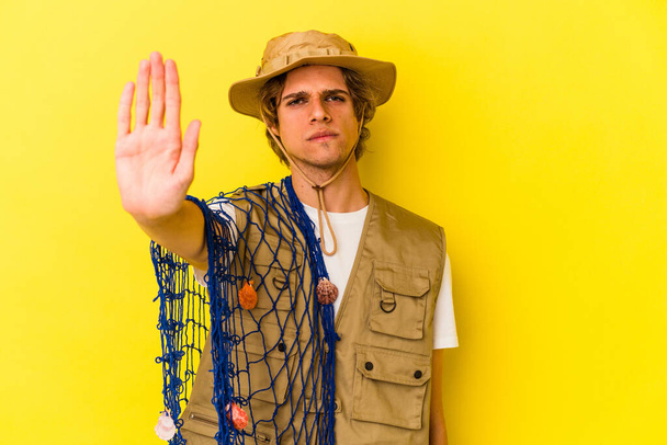 Young fisherman with makeup holding a net isolated on yellow background  standing with outstretched hand showing stop sign, preventing you. - Photo, Image