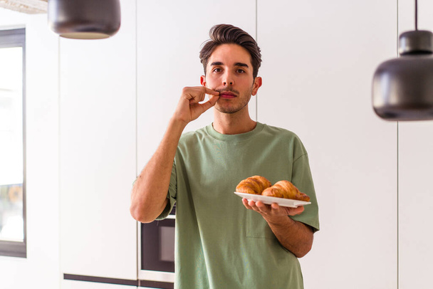 Jeune homme métis mangeant croissant dans une cuisine le matin avec les doigts sur les lèvres gardant un secret. - Photo, image