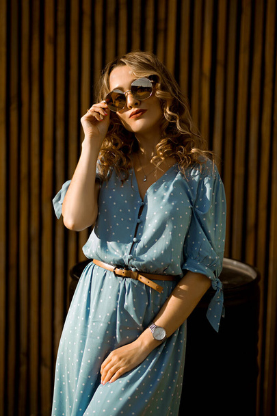 portrait of a young caucasian curly-haired woman in a long blue dress, european lady in glasses on a summer street - Foto, Bild