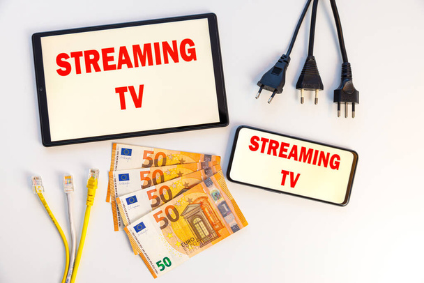 Tabletták és okostelefonok, amelyek a kijelzőn a "streaming tv" feliratot, bankjegyeket, adatkábeleket, pénzt és elektromos csatlakozókat mutatják. A streaming TV programok költségei és fogyasztása.  - Fotó, kép