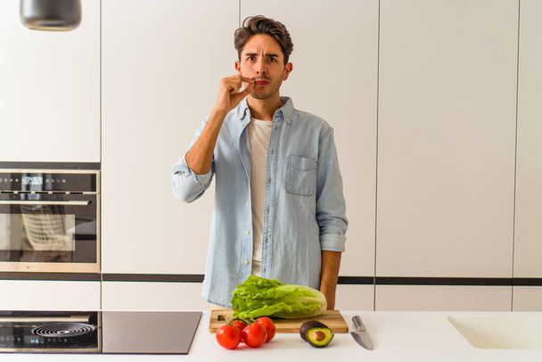 jonge gemengde ras man voorbereiding van een salade voor de lunch met vingers op de lippen houden van een geheim. - Foto, afbeelding