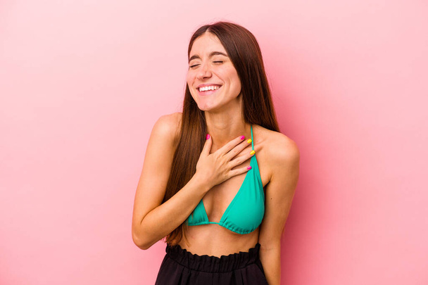 Joven mujer caucásica con bikini aislado sobre fondo rosa se ríe en voz alta manteniendo la mano en el pecho. - Foto, imagen