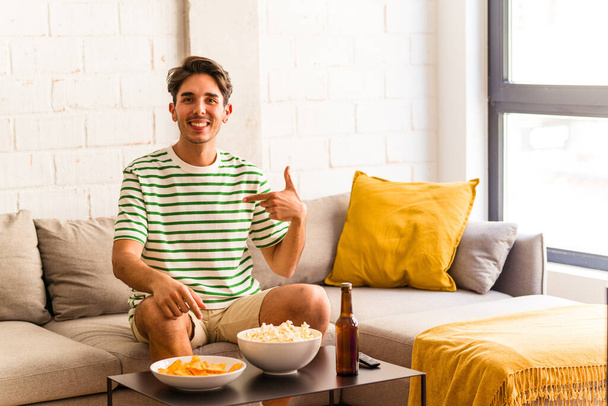 Молодой человек смешанной расы ест попкорны сидя на диване человек указывая вручную на рубашку копировать пространство, гордый и уверенный - Фото, изображение