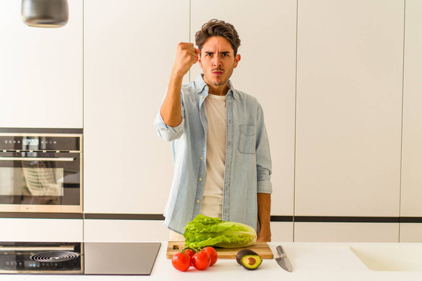 Young mixed race man preparing a salad for lunch showing fist to camera, aggressive facial expression. - Photo, Image