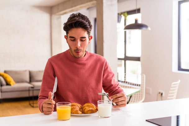 Fiatal vegyes fajú férfi eszik croissant a konyhában reggel - Fotó, kép