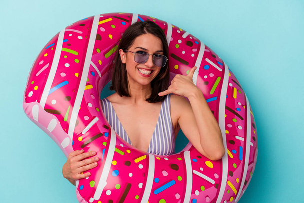 Young caucasian woman with inflatable donut isolated on blue background showing a mobile phone call gesture with fingers. - Foto, afbeelding
