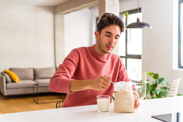Young mixed race man eating oatmeal and milk for breakfast in his kitchen - 写真・画像