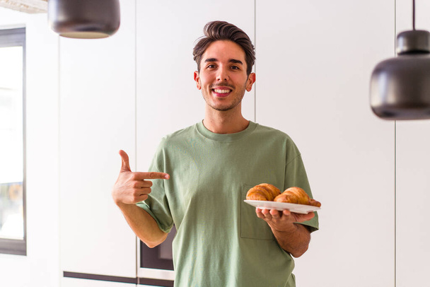 Jeune homme métis mangeant croissant dans une cuisine le matin personne pointant à la main vers un espace de copie de chemise, fier et confiant - Photo, image