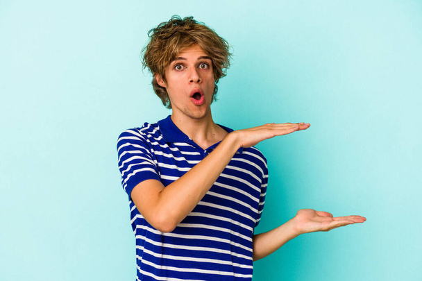 Young caucasian man with make up isolated on blue background  shocked and amazed holding a copy space between hands. - Photo, Image