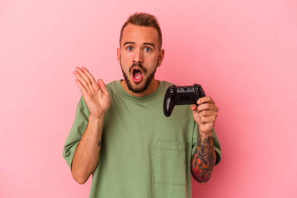 Jovem caucasiano com tatuagens segurando controlador de jogo isolado no fundo rosa surpreso e chocado. - Foto, Imagem
