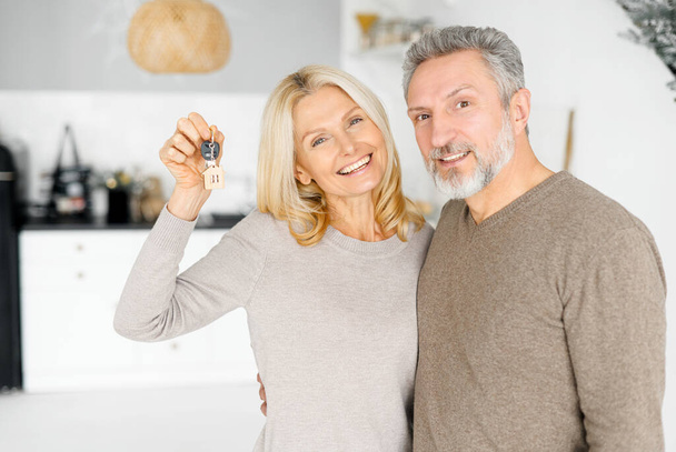 Charming middle-aged wife holding home keys standing in hug of her husband - Foto, Bild
