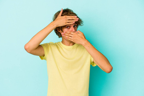 Young caucasian man with make up isolated on blue background  blink at the camera through fingers, embarrassed covering face. - Fotoğraf, Görsel