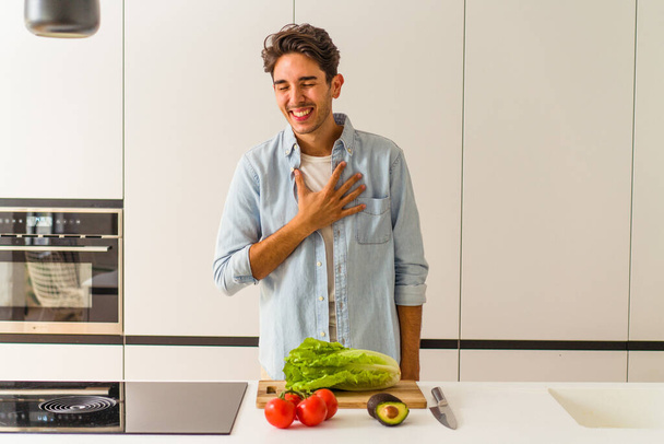 Молодий змішаний чоловік готує салат на обід сміється голосно тримаючи руку на грудях
. - Фото, зображення