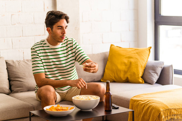 Молодий змішаний чоловік їсть попкорн, сидячи на дивані
 - Фото, зображення