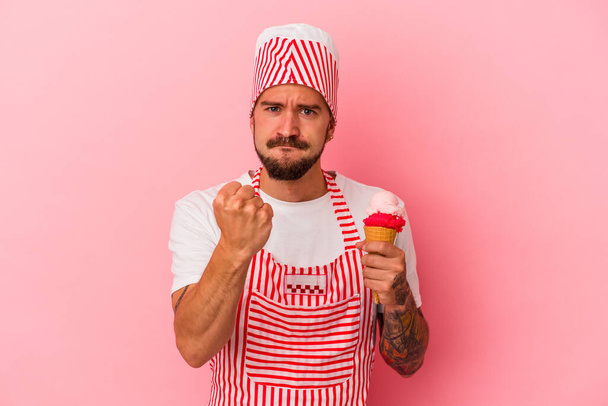Jeune homme de machine à glace caucasien avec des tatouages tenant la crème glacée isolée sur fond rose montrant poing à caméra, expression faciale agressive. - Photo, image