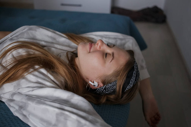 Молодая 20-летняя девушка с повязкой на голове и длинными светлыми волосами лежит на кровати и слушает музыку в наушниках - Фото, изображение