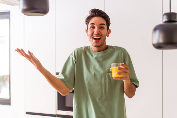 Jonge gemengde ras man drinken sinaasappelsap in zijn keuken ontvangen van een aangename verrassing, opgewonden en het verhogen van de handen. - Foto, afbeelding