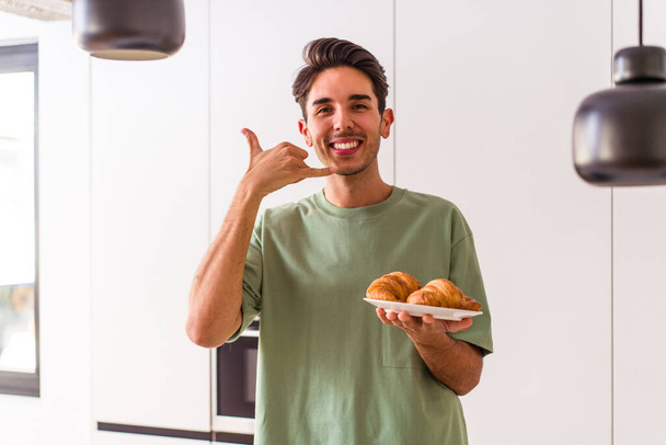 Jeune homme métis mangeant croissant dans une cuisine le matin montrant un geste d'appel téléphonique mobile avec les doigts. - Photo, image