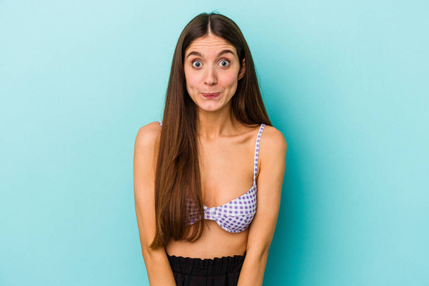 Young caucasian woman wearing bikini isolated on blue background shrugs shoulders and open eyes confused. - Photo, Image